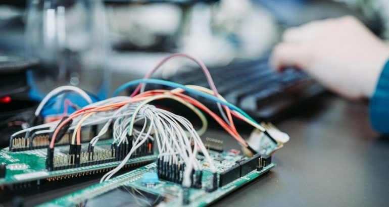 Profissional trabalhando em computador próximo a uma placa eletrônica, ilustrando análise técnica de softwares